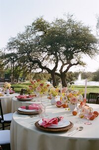Spring wedding reception tablescape on film photo at Ma Maison in Dripping Springs, Texas, featuring citrus inspired florals, pink napkins, and a view of the oak trees and fountain.