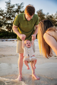 Family of three. Dad is holding onto the toddlers arms as he lifts his legs so he doesn't touch the water. He is full of smiles. Mom is facing away from the camera looking at her child who is looking at her. She is bent over so she is at the same height.