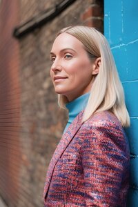 JO WOOD BLONDE WOMAN IN BLUE POLO NEXK JUMPER AND PINK JACKET STOOD NEXT TO A BLUE WALL SIDE PROFILE