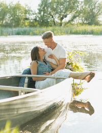 A man and a woman are sitting candidly in a rowboat. The girl is dangling her feet off the side and looking up at her fiance, smiling.