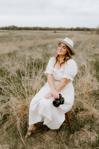 woman sitting in a field wearing a dress and hat and holding a camera