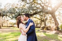 Bride and groom sharing a kiss under moss-draped oak trees in a sunny Savannah park during their elopement.