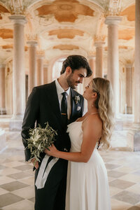 In einer hellen Säulenhalle steht ein Hochzeitspaar eng beieinander und blickt sich tief in die Augen.