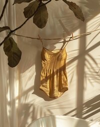 Golden satin camisole hanging on a branch in soft sunlight, with plant shadows cast on the wall — warm, minimal and serene aesthetic.