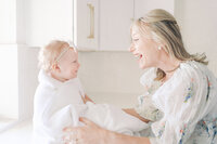 mom in white floral dress looking at one year old daughter smiling at each other. Daughter wrapped in white towel wearing a white bow