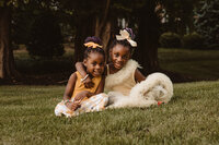 Two siblings sitting in the grass with their arms around each other smiling 