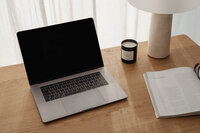 A minimalist wooden desk setup featuring an open laptop, a beige lamp, an open book, and a candle. Soft natural light filters through white net curtains, creating a calm and inspiring workspace aesthetic.