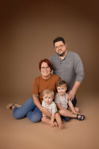A family of four poses against a brown backdrop. The mother, with short red hair and glasses, sits on the floor with their two young children in her lap. The father, wearing glasses and a grey shirt, stands behind them, smiling. Capturing this moment is a testament to Jacksonville family photography.