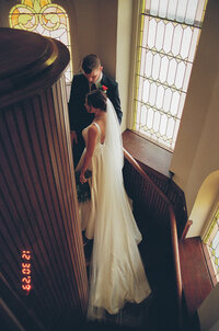 Bride and groom standing in a wooden curved stairway in a church surrounded by stained glass windows.