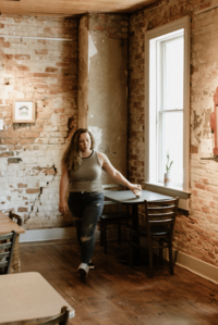 A woman in a gray tank top and jeans walks through a rustic cafe with exposed brick walls, wooden floors, and sunlight streaming in—perhaps dreaming of how to become a white label designer.