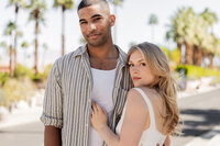 Engaged couple standing closely in the Palm Springs sun, surrounded by palm trees and desert textures, captured in a soft, romantic moment.