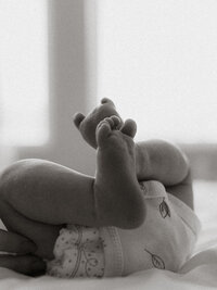 Black and white photo of newborn baby’s feet lifted gently in the air during a quiet in-home session, capturing the tenderness of early days.