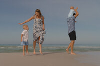 A photo of a young son and a mother playfully jumping in the air. The father has their youngest son on his shoulders while he is walking. They are on the beach in 30a, Florida. Storytelling Family Photographer Santa Rosa Beach, FL. 