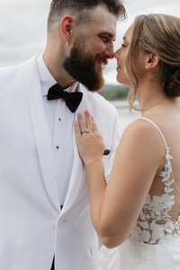 A bride and groom kiss by a scenic pond, captured beautifully by a New England Photographer. The bride stuns in her lace gown, the groom charming in his white shirt and suspenders. He holds a vibrant bouquet of red, blue, and yellow flowers against the backdrop of lush trees and greenery.
