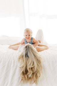Portrait of a woman wearing white pants lying in bed while holding a smiling baby wearing blue  in a bright and airy room