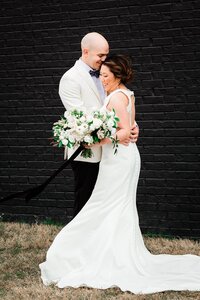 Asian bride and groom wearing white hugging against a black backdrop with large white bouquet