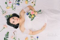 Pregnant woman lying on a white sheet surrounded by colorful flowers, holding a bouquet of pink, yellow, and blue blooms while wearing an off-shoulder lace dress.