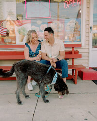 A couple sitting on a bench outside an ice cream shop with their dog in front of them 