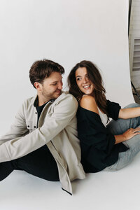 Wedding and elopement photographer and videographer sit back to back, smiling at each other