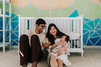 mom holding newborn daughter while husband sits next to them. They are laughing and sitting against a crib with a geometric rainbow nursery mural