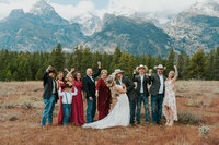 A wedding party consisting of the bride, groom, and 9 other family members posing in front of the Tetons. The bride and groom are about to kiss while everyone else is cheering.