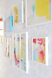 A colorful gallery wall of paintings in white frames.