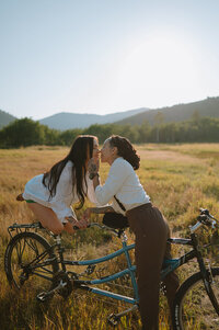 Balancing on a bike, a woman kisses her girlfriend who is standing next to her. They're in beautiful grass field in Swan Valley, Idaho.