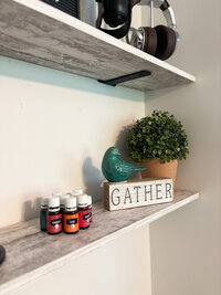 Wooden shelf featuring essential oils, greenery, and a decorative "gather" sign with cozy vibes.