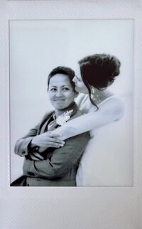 Photo of two brides in black and white, smiling at the camera, with one wearing a dress and one wearing a suit, standing on the beach in Cape May