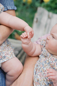 twin babies with pudgy rolled arms reach to hold hands wearing floral dresses by Portland Family Photographer Emilie Phillipson Photography. 