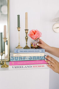 Marissa Martinez's hands setting a flower in a vase on top of a stack of colorful books next to gold candlesticks