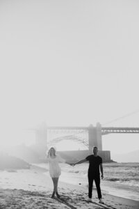 Couple walking at beach with Golden Gate Bridge in background at sunset golden hour