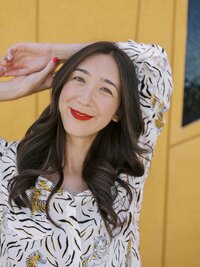 Woman smiling in a patterned blouse posing with arms raised in front of a mustard yellow wall — NYC brand photography shoot.