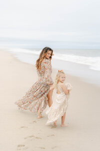 Mother with long brown hair and flowy dress walks the beach in Delaware with her daughter during a family photo session