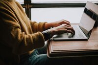 A person in a yellow sweater sits at a table in front of a window and types on a laptop