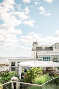 A view of the Cliff House Maine’s bright white exterior and waterfront pergola, with lush greenery and a sparkling ocean backdrop under a sunny sky.