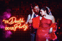 A bride in a bright red dress embraces her husband next to a sign that says "Til death do us party"