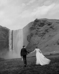 Black and white photo of a couple walking hand-in-hand toward Skógafoss waterfall in Iceland during their adventurous elopement — the bride’s flowing gown trailing behind as the mist rises from the falls, photographed by Destination Elopement Photographer