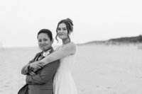 Photo of two brides in black and white, smiling at the camera, with one wearing a dress and one wearing a suit, standing on the beach in Cape May