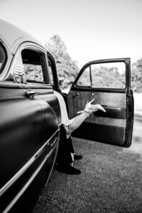 Black and white photo of a bride playfully kicking up her heels while stepping out of a vintage car, capturing a timeless and elegant elopement moment.