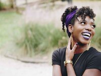 A young Black feminine presenting person looks over their left shoulder, past the camera, while smiling widely. They are wearing a dark purple headband around their curly hair, as well as dark purple lipstick and a black t-shirt.