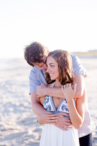 Portrait of a couple on the beach. The boy is kissing the girl and the girl is looking out to the ocean. She is wearing a white maxi dress and they are hugging.