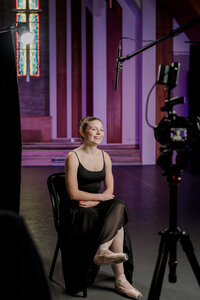 A smiling female ballerina in a black leotard and skirt sits in a chair on a backlit stage in front of a camera.