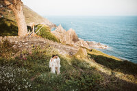 A wedding couple on the coast of looe england hugging with the cliffs and ocean behind them while standing in a flower field.
