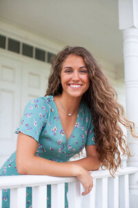 A lovely brunette wavy haired girl in a blue floral dress leaning over a white porch railing. Captured by Springfield, MO photographer Dynae Levingston.