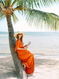 Kilee in an orange dress and straw hat sitting on a palm tree with a book in front of the ocean