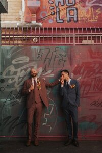 Two individuals, each dressed in stylish suits—one in maroon and the other in navy—pose affectionately against a wall covered in vibrant graffiti. One person holds the other's hand close to their lips in a tender gesture.