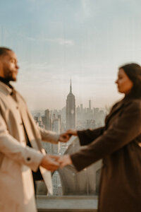 Couple hold hands on Top of the Rock in NYC overlooking the empire state building.