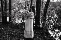 Black and white photo of a mom holding a toddler while they smile at each other in a forested area.