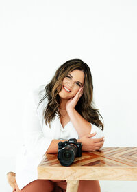 Adriana Montoya Photo sitting at a small table. One arm is propped against the side of her head, holding her head up. Her other arm is touching the elbow of her other arm. On the table in front of her is her camera.